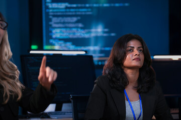 Two female developers, one Indian and one Caucasian, in a cybersecurity command center SOC. They are looking up, analyzing AI code and system data during a technical briefing at night.