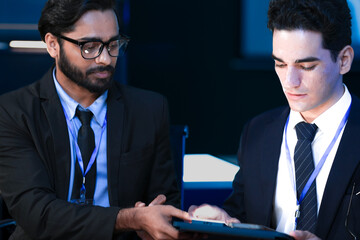 An Indian developer holds a tablet while a Caucasian engineer interacts with the screen. They are analyzing AI data or a cybersecurity system in a SOC.