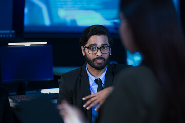 An Indian male data scientist speaks during a late-night technical briefing in a SOC. His female engineer colleague listens while analyzing AI or cybersecurity data on the screens.