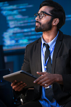 An Indian male software engineer or data scientist holds a tablet in a command center. He is asking a question about the AI code or cybersecurity data shown on the large screen.
