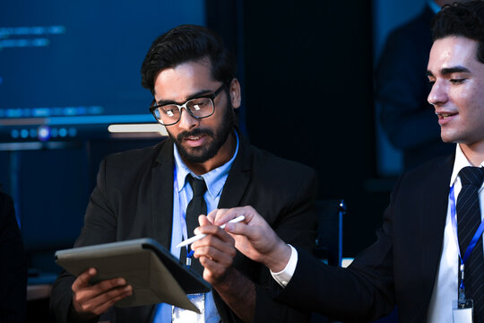 An Indian man with glasses uses a tablet to explain a project to his smiling Caucasian male colleague. They are collaborating in a modern high tech office.