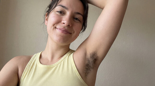Confident young woman with natural armpit hair smiling in casual yellow top against neutral indoor wall background