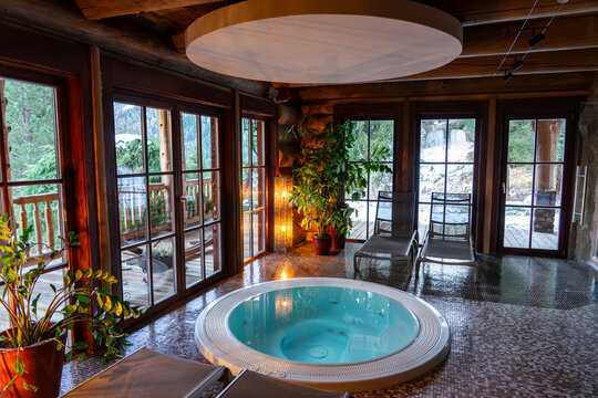 Indoor jacuzzi in a warm wooden cabin hotel with large windows overlooking snowy forest .