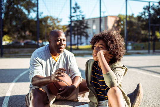Grandfather and grandson resting on outdoor basketball court, thoughtful