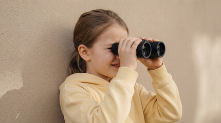 Curious child in yellow hoodie using binoculars to observe something in the distance while leaning against a beige outdoor wall
