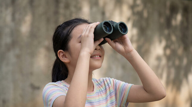 Curious young girl observing distant scenery with binoculars outdoors against neutral wall in soft natural daylight
