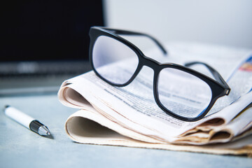 A pile of newspapers ,glasses and computer on the table.