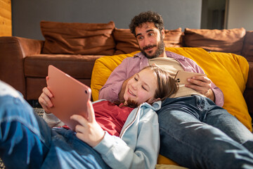 Adult father and child daughter relaxing with tablet on living room sofa, happy