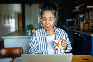Mature woman focused on laptop with coffee in kitchen