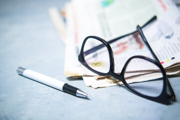 A pile of newspapers ,glasses and pen on the table.