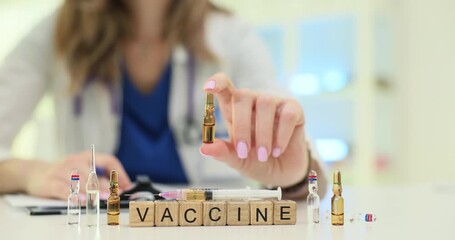 Woman doctor in clean white medical coat shows small transparent ampoule with medication. Miniature wooden cubes on table form word Vaccine