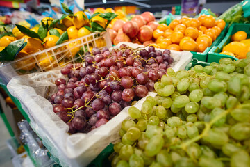 Fresh fruits are displayed in baskets at a grocery store. The scene features green grapes, oranges.