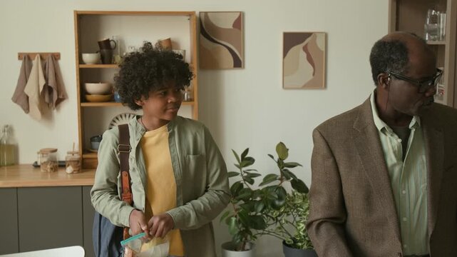 Senior African American man kissing goodbye two young grandsons helping kids leaving for school with healthy snacks, family morning routine in kitchen