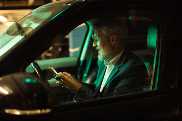 Mature businessman focused on smartphone in car at night