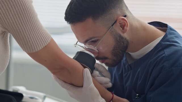 Chest up slowmo shot of concentrated young Middle-Eastern male dermatologist adjusting eyeglasses and inspecting skin on arm of unrecognizable female patient using dermatoscope in clinic