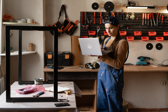 Female small business owner standing in repair workshop holding laptop for SME management tasks