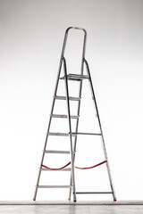 A tall metal step ladder is positioned upright against a neutral backdrop. The ladder features several steps and safety straps, reflecting a simple, functional design often used in various tasks
