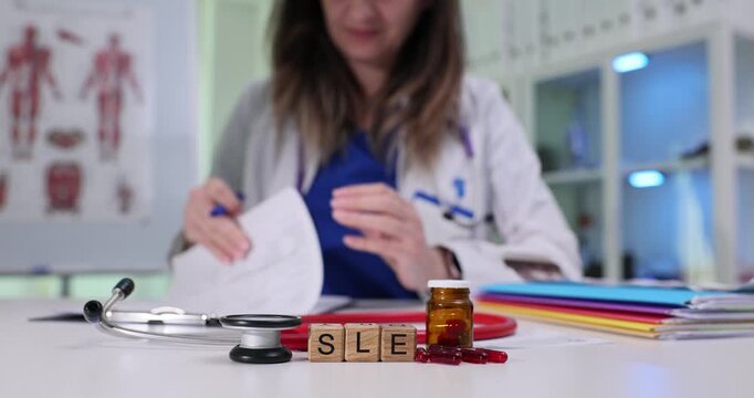 Concentrated woman doctor examines medical history of patient and makes detailed notes. Wooden cubes arranged on table form abbreviation SLE