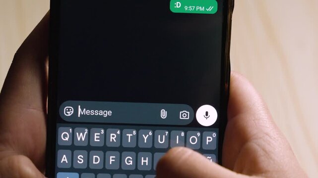 Close-up view of hands holding a smartphone while typing the emoticon &ldquo;:D&rdquo; in a messaging app, with a thumb hovering over the send button on a dark-themed keyboard.