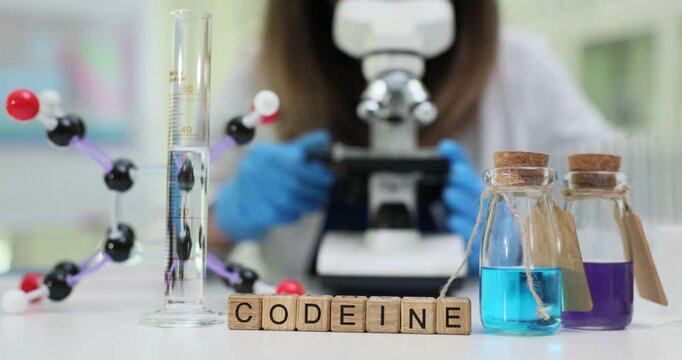 Woman chemist adjusts fine focus knob of microscope to study Codeine. Wooden cubes on laboratory table precisely arranged to form word Codeine