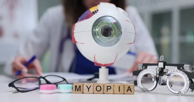 Artificial eyeball model stands on desk and wooden cubes form word Myopia. Ophthalmologist writes recommendations for patient corrective lenses