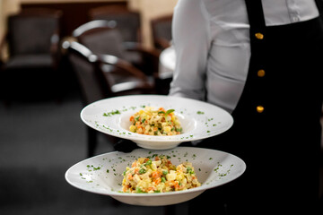 A server in a restaurant presents two plates of creamy Olivier salad, garnished with herbs, ready to be served to diners