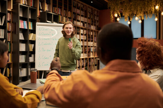 Young man standing near flip chart teaching diverse group of young people in library classroom setting, students listening attentively and taking notes during language lesson