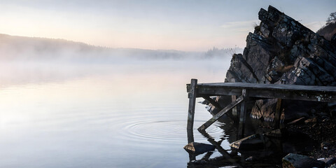 Serene foggy sunrise over calm lake, with ripples reflecting in the water and old wooden pier next to steep, moss-covered rock formation.