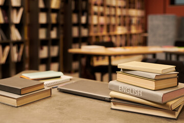 Stack of books including English language textbook on table in library with shelves full of books in background
