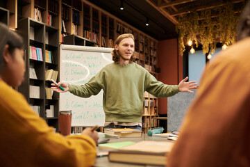 Young man standing in front of whiteboard teaching diverse group of students in library classroom, gesturing with arms open while explaining lesson content