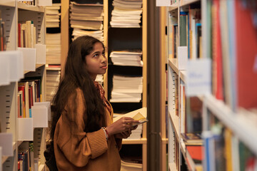Teenage Asian girl standing between bookshelves holding open book, looking up thoughtfully, surrounded by stacks of books in library setting, long dark hair