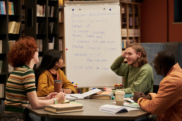 Group of diverse young people sitting around table engaging in language learning activity, man leading discussion, books and notebooks scattered, flip chart displaying French phrases