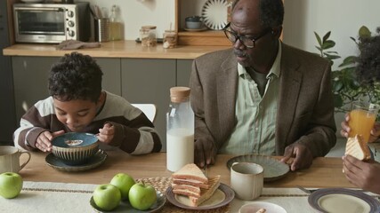 Elderly Black man caring for preteen and teenage grandsons while having cereal flakes and sandwiches for breakfast at kitchen table, family morning routine - Powered by Adobe