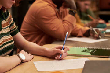 Young woman writing on paper at desk beside Black young man taking notes in classroom setting, both...