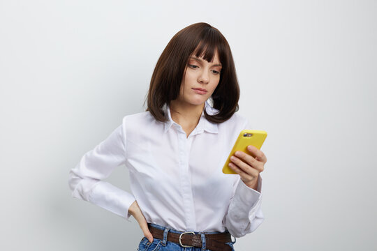 A stylish woman checks her smartphone while standing against a clean, light background, wearing a white shirt and jeans. The casual scene conveys focus, modern work life, and personal communication