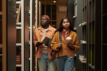 Young Black man holding book and young Asian woman carrying coffee cup and backpack walking between bookshelves in library, both looking ahead, studying together