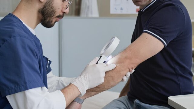 Cropped shot of young Middle Eastern male doctor inspecting male patient with skin rash on elbow using magnifying glass during consultation in modern dermatology clinic