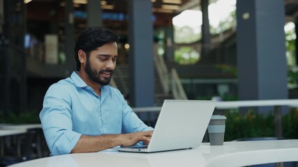 Portrait of smiling Indian businessman controls cryptocurrency rates on laptop in urban cafe. Young man investor works on notebook computer at table outdoors. Financial technology - Powered by Adobe