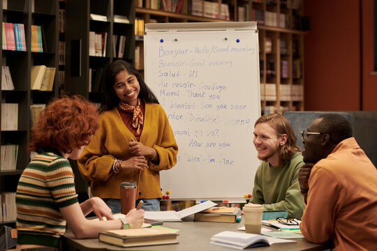 Multiethnic group of young people sitting around table listening to smiling young Asian woman presenting during language class in library