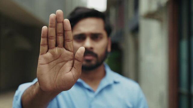 Portrait of serious Indian man raises arm showing Stop sign on narrow street closeup. Bearded male in blue shirt with No gesture expresses disagreement in old town district