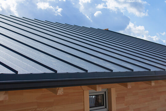 Dark gray standing seam metal roof and black gutter system on a building with natural wooden log walls for exterior building materials. Residential construction and home improvement.