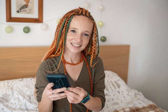 Cheerful woman with braided red hair and freckles holding a smartphone and smiling at the camera.