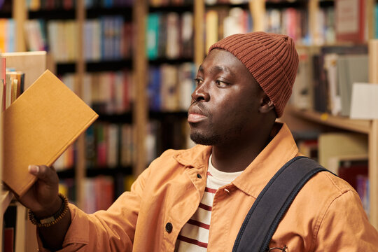 Young Black man browsing bookshelves in library, reaching for hardcover book with focused expression, wearing casual clothing and carrying shoulder bag, surrounded by books