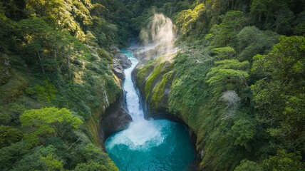 Aerial view of a stunning turquoise waterfall cascading into a deep pool surrounded by lush, dense green tropical rainforest canopy under bright sunlight