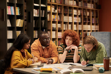 Group of diverse young people studying together at table in library, multiethnic men and women collaborating on academic project, reading books, taking notes, discussing assignments
