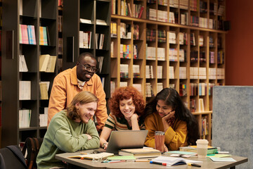 Group of diverse young people collaborating around laptop in library, man, Black man, woman, Asian woman studying together, smiling and engaged in teamwork