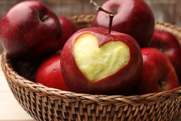 Ripe apple with carved heart and whole ones in wicker basket on light table, closeup