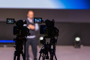 Cameras set up focus on blurred presenter delivering talk while audience unseen. Behind scenes shot...