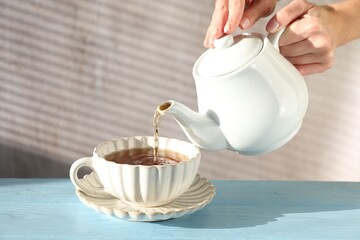 Woman pouring hot tea into cup from teapot at light blue wooden table, closeup