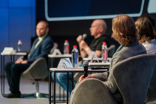 Panel discussion at conference features speakers seated in soft chairs microphones on table water bottles name cards. Professional focused atmosphere fosters collaboration learning inspiration 
